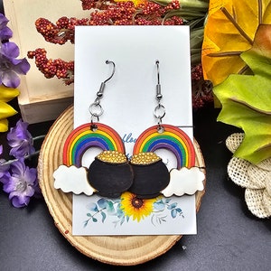 Könnte beinhalten: Ein Paar Regenbogen- und Goldtopf-Ohrringe. Die Ohrringe sind aus Holz und zeigen einen Regenbogen mit einem Topf Gold am Ende. Die Ohrringe hängen an silbernen Haken.