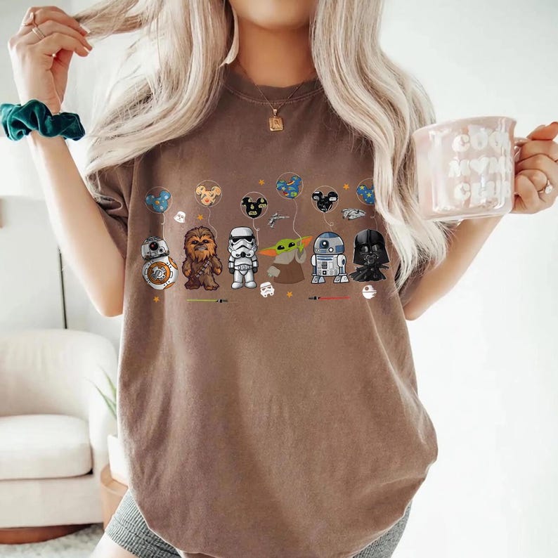 Op de afbeelding: Bruin t-shirt met cartoon illustraties van Star Wars personages, waaronder BB-8, Chewbacca, een Stormtrooper, Grogu, R2-D2 en Darth Vader. Het ontwerp bevat ook ballonnen en lichtzwaarden met personage thema's.