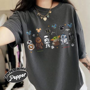 Op de afbeelding: Donkergrijs Comfort Colors T-shirt met een Star Wars-karakterafbeelding. De afbeelding bevat BB-8, Chewbacca, een Stormtrooper, Grogu, R2-D2 en Darth Vader, elk met een ballon. De tekst "Pepper" staat onder de woorden "Comfort Colors".