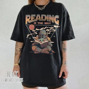 May include: Black t-shirt with a graphic featuring a Mandalorian character reading to Grogu. The text "READING IS THE WAY" is above the illustration. The words "BLACK COMFORT COLORS" are printed at the bottom.