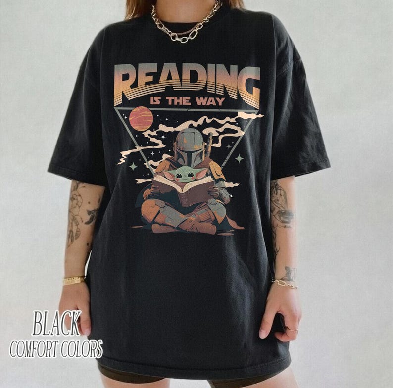May include: Black t-shirt with a graphic featuring a Mandalorian character reading to Grogu. The text "READING IS THE WAY" is above the illustration. The words "BLACK COMFORT COLORS" are printed at the bottom.