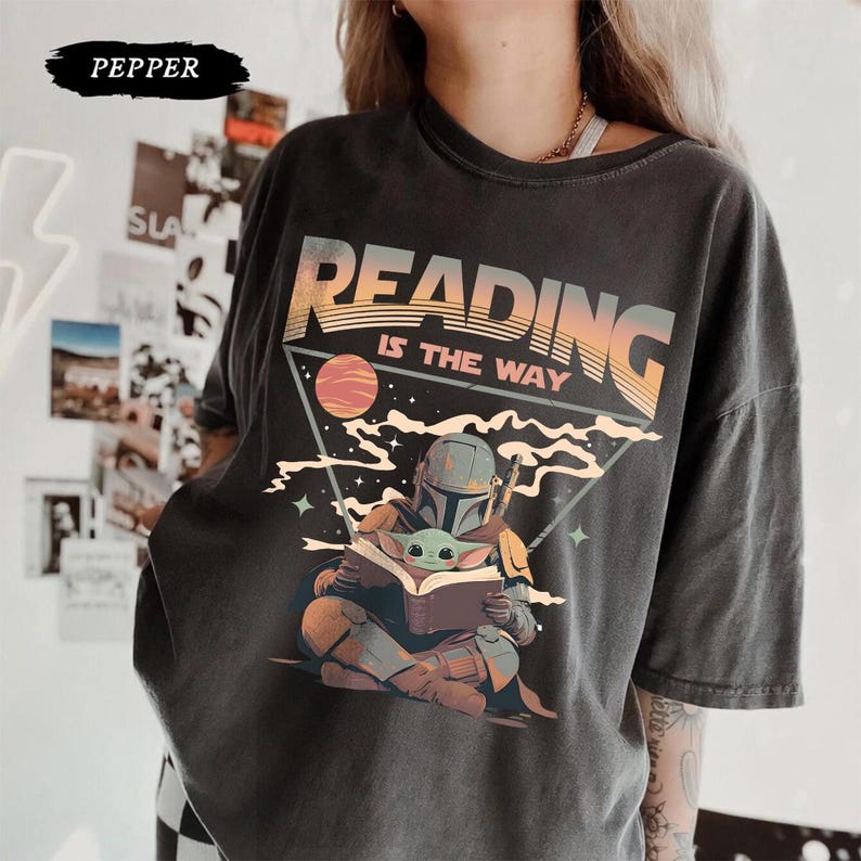 May include: Dark gray t-shirt with the text "READING IS THE WAY" above an illustration of a Mandalorian character reading a book to Grogu. The design features a planet and stars within a triangle.