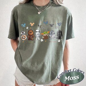 Op de afbeelding: Olijfgroen T-shirt met Star Wars-personages die Mickey Mouse-ballonnen vasthouden. Personages zijn onder meer BB-8, Chewbacca, Stormtrooper, Grogu, R2-D2 en Darth Vader. Het shirt heeft de woorden "Comfort Colors Moss" rechtsonder.