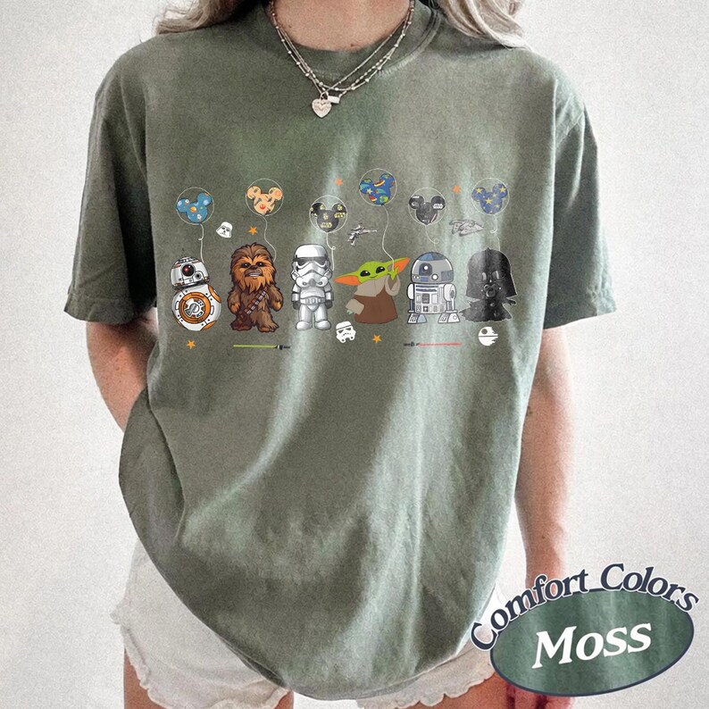 Op de afbeelding: Olijfgroen T-shirt met Star Wars-personages die Mickey Mouse-ballonnen vasthouden. Personages zijn onder meer BB-8, Chewbacca, Stormtrooper, Grogu, R2-D2 en Darth Vader. Het shirt heeft de woorden "Comfort Colors Moss" rechtsonder.