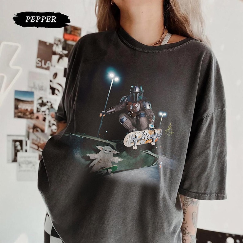 May include: Dark gray t-shirt featuring a graphic of The Mandalorian skateboarding, with Grogu on a skateboard below. The image is set in a skate park at night. The word "PEPPER" is printed at the top.