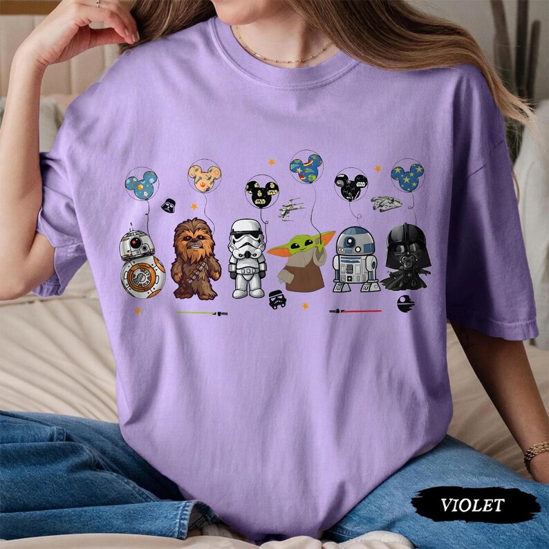 Op de afbeelding: Een lichtpaars t-shirt met Star Wars-personages en Mickey Mouse-vormige ballonnen. Het ontwerp bevat BB-8, Chewbacca, een Stormtrooper, Grogu, R2-D2 en Darth Vader. Het woord "VIOLET" staat onderaan afgedrukt.