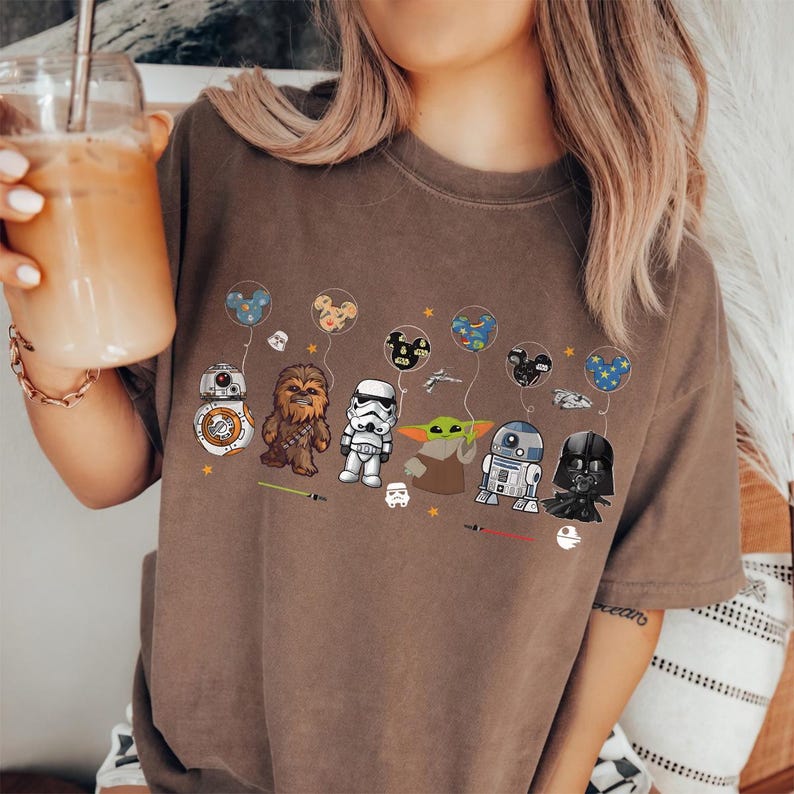 Op de afbeelding: Bruin t-shirt met Star Wars-personages, waaronder BB-8, Chewbacca, een Stormtrooper, Grogu, R2-D2 en Darth Vader. Elk personage houdt een ballon vast met een Mickey Mouse-hoofdontwerp. Het t-shirt heeft een relaxte pasvorm.
