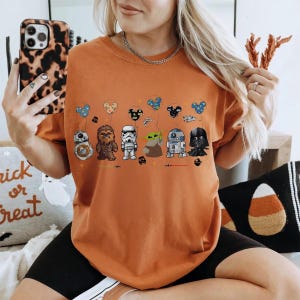 Op de afbeelding: Oranje T-shirt met Star Wars-personages als ballonnen. Het ontwerp omvat BB-8, Chewbacca, een Stormtrooper, Grogu, R2-D2 en Darth Vader. De ballonnen hebben de vorm van Mickey Mouse-hoofden. Het shirt is een casual stijl.