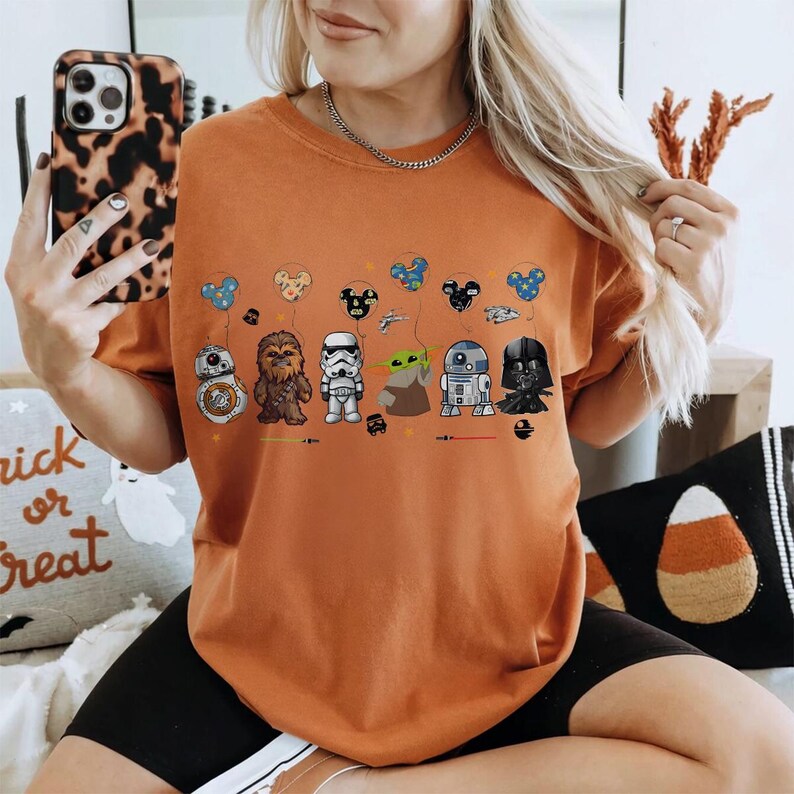 Op de afbeelding: Oranje T-shirt met Star Wars-personages als ballonnen. Het ontwerp omvat BB-8, Chewbacca, een Stormtrooper, Grogu, R2-D2 en Darth Vader. De ballonnen hebben de vorm van Mickey Mouse-hoofden. Het shirt is een casual stijl.