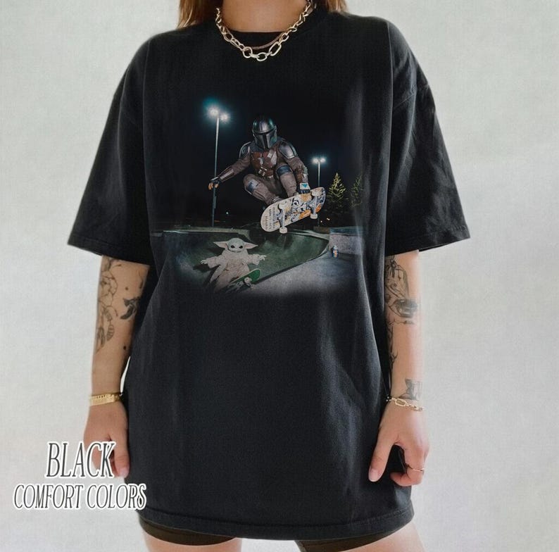 May include: Black comfort colors t-shirt featuring a graphic of a character from The Mandalorian skateboarding in a skate park, with a smaller character also skateboarding. The text "BLACK COMFORT COLORS" is printed at the bottom.