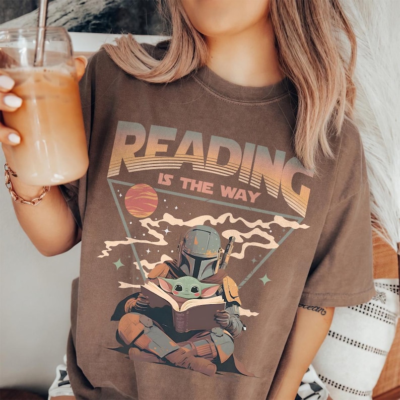 May include: Brown t-shirt with the text "READING IS THE WAY" in an orange and tan retro font. The graphic features a Mandalorian character and Grogu reading a book, with a planet and stars in the background.