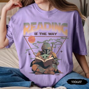 May include: Violet short-sleeved t-shirt with the text "READING IS THE WAY" in a gradient design. The graphic features a Mandalorian character reading a book with Grogu, set against a triangle and planet.