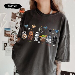 Op de afbeelding: Donkergrijs T-shirt met een Star Wars-afbeelding. Het ontwerp toont cartoonpersonages zoals BB-8, Chewbacca, een Stormtrooper, Grogu, R2-D2 en Darth Vader, elk met een ballon. Het woord "PEPPER" staat bovenaan.