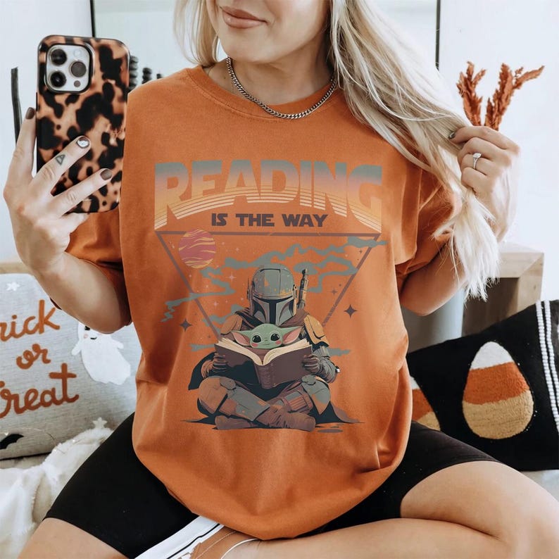 May include: Orange t-shirt with the text "READING IS THE WAY" above an illustration of a Mandalorian character and Grogu reading a book. The design includes a triangle, planets, and stars. The shirt is a comfortable fit.
