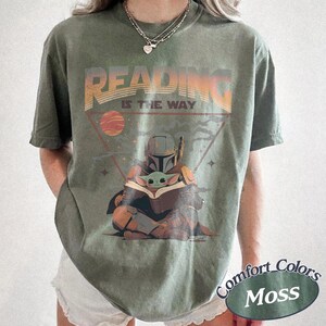 May include: Olive green t-shirt with the text "READING IS THE WAY" above an illustration of The Mandalorian and Grogu reading a book. The design includes a planet and stars within a triangle. The shirt has a Comfort Colors Moss label.