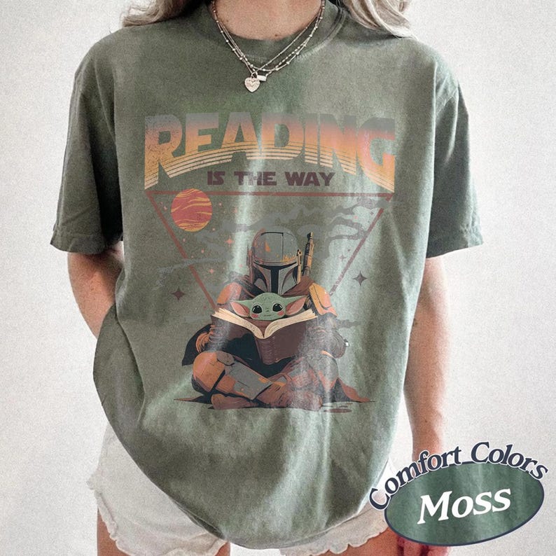 May include: Olive green t-shirt with the text "READING IS THE WAY" above an illustration of The Mandalorian and Grogu reading a book. The design includes a planet and stars within a triangle. The shirt has a Comfort Colors Moss label.