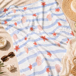 May include: A beach towel with blue and white stripes, adorned with red starfish and pink seashell illustrations. The towel is laid out on sand, with a straw hat, sunglasses, and a small bowl nearby.
