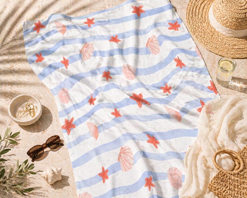 May include: A beach towel with blue and white stripes, adorned with red starfish and pink seashell illustrations. The towel is laid out on sand, with a straw hat, sunglasses, and a small bowl nearby.