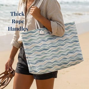 May include: A large tote bag with a wave pattern in shades of blue, green, and beige. The bag has thick rope handles and the text "Thick Rope Handles" is visible. The bag is being held on a beach.