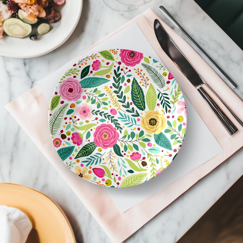 May include: A white ceramic plate with a floral design featuring pink roses, yellow flowers, and green leaves. The plate is set on a pink napkin with silverware and a marble table in the background. The plate is approximately 10 inches in diameter.