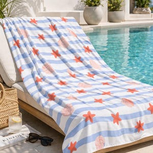 May include: A beach towel with a blue and white striped pattern, adorned with coral-colored starfish and seashell designs. The towel is draped over a wooden chaise lounge near a pool, with a straw tote bag and sunglasses nearby.