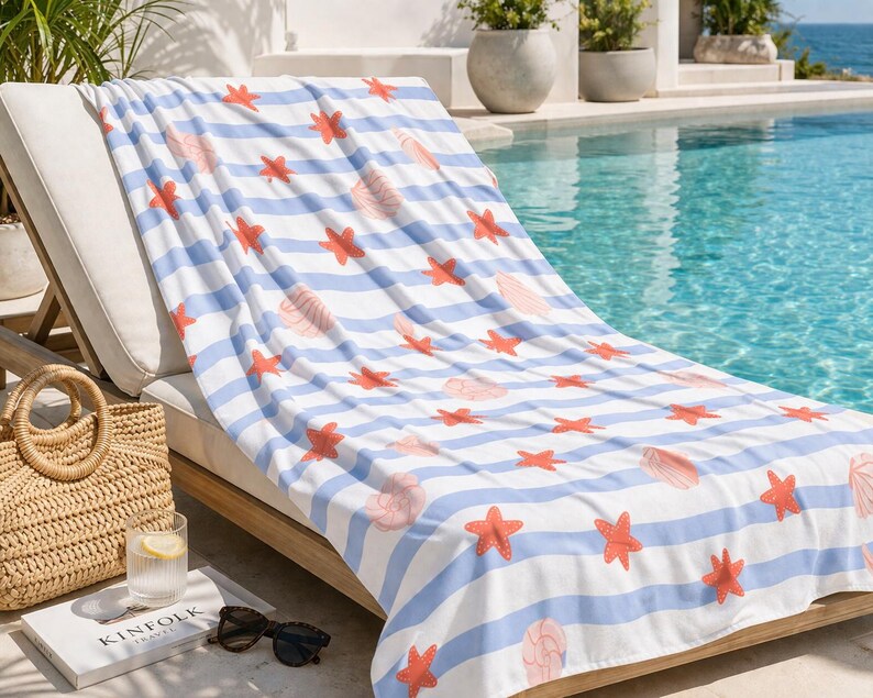 May include: A beach towel with a blue and white striped pattern, adorned with coral-colored starfish and seashell designs. The towel is draped over a wooden chaise lounge near a pool, with a straw tote bag and sunglasses nearby.