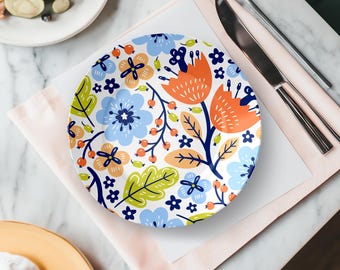 May include: A round white plate with a colorful floral design featuring orange, blue, and green flowers and leaves. The plate is set on a pink napkin with silverware and a partially visible plate of food.