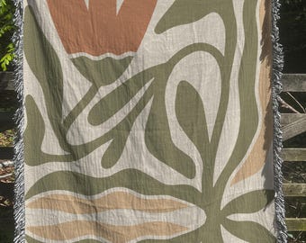 May include: A woven throw blanket with a large abstract floral design in shades of green, brown, and beige. The blanket has a fringe border.