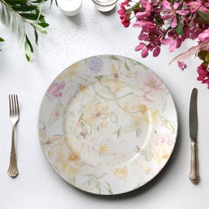 May include: A white ceramic dinner plate with a floral design in soft yellow, pink, and lavender hues. A silver fork and knife are placed on either side of the plate. The plate is surrounded by greenery and pink flowers.