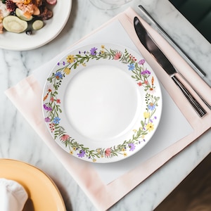 May include: A white ceramic plate with a floral border featuring colorful flowers and green leaves. The plate is set on a pink napkin with silverware and a marble table in the background. A second plate with food is visible.