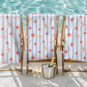 May include: Three wooden beach chairs with white and blue striped towels adorned with red starfish and seashell patterns. A silver bucket with a champagne bottle and two glasses of champagne are in front of the chairs.