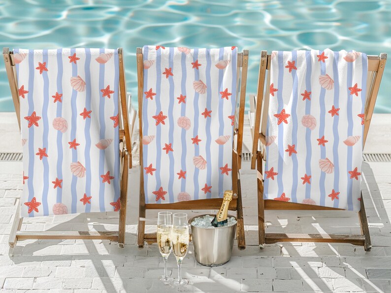 May include: Three wooden beach chairs with white and blue striped towels adorned with red starfish and seashell patterns. A silver bucket with a champagne bottle and two glasses of champagne are in front of the chairs.