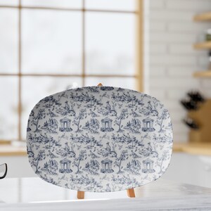 May include: Oval serving platter with a blue and white toile pattern. The design features classical architectural elements, trees, and figures. The platter is displayed on a small wooden easel, suitable for serving appetizers or desserts.
