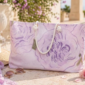 May include: A large, rectangular tote bag with a floral design in shades of lavender and purple. The bag has white rope handles and is set on a stone surface with a straw hat, sunglasses, and a scarf nearby. A pink flower and pool are in the background.