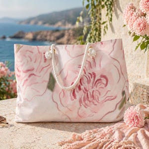 May include: A large, light pink tote bag with a watercolor floral design. The bag has white rope handles and is sitting on a stone surface. Sunglasses and a pink scarf are next to the bag. The background shows a coastal view.