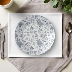 May include: A round white plate with a blue floral pattern, featuring various flowers and leaves. The plate is set on a white square napkin, with a gray napkin underneath. A spoon and a knife are visible.