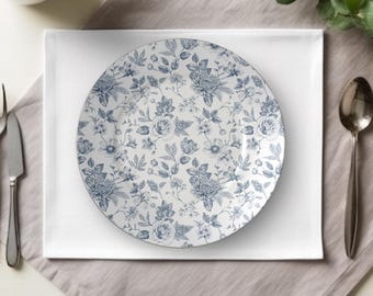 May include: A round white plate with a blue floral pattern, featuring various flowers and leaves. The plate is set on a white square napkin, with a gray napkin underneath. A spoon and a knife are visible.
