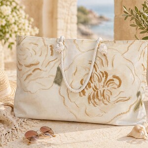 May include: A large cream-colored tote bag with a floral design in shades of brown and green. The bag has rope handles and is sitting on a stone surface. A straw hat and sunglasses are nearby.