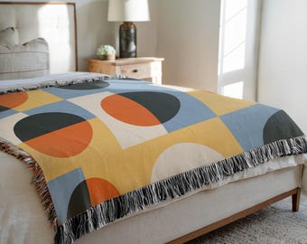 May include: A woven throw blanket with a geometric pattern of circles and squares in shades of yellow, blue, orange, black, and white. The blanket has a fringe trim and is displayed on a bed. This blanket is perfect for adding a pop of color to any room.