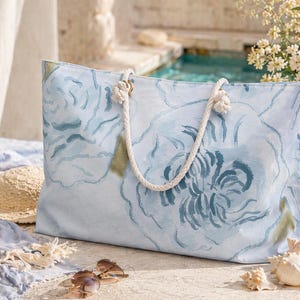 May include: A large, rectangular tote bag with a light blue floral design. The bag has white rope handles and gold-colored metal rings. The background includes a straw hat, sunglasses, and seashells, suggesting a beach or vacation theme.