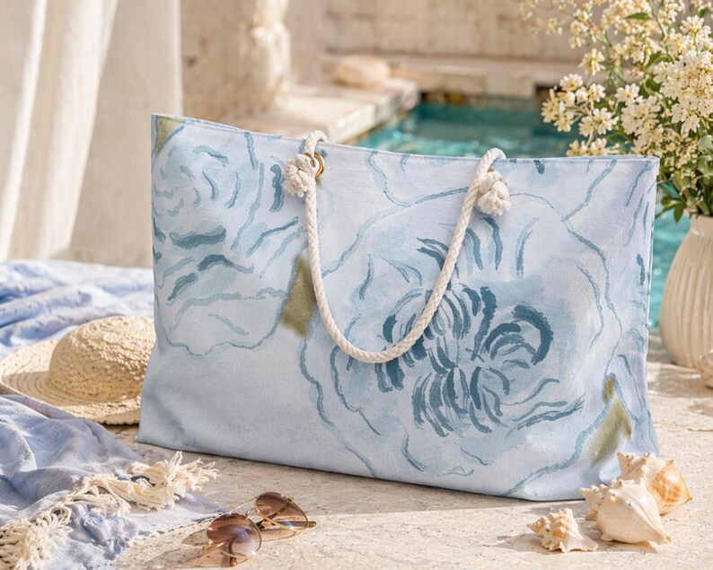 May include: A large, rectangular tote bag with a light blue floral design. The bag has white rope handles and gold-colored metal rings. The background includes a straw hat, sunglasses, and seashells, suggesting a beach or vacation theme.