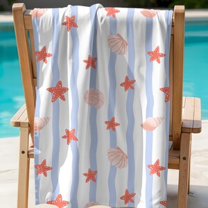 May include: Beach towel with a pattern of red starfish and seashells on a white background with light blue vertical stripes. The towel is draped over a wooden chair, with a pool in the background.