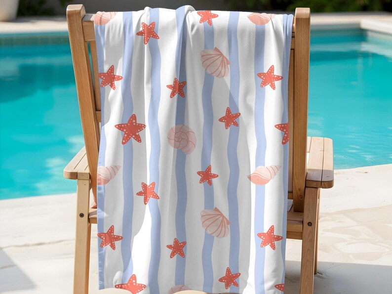 May include: Beach towel with a pattern of red starfish and seashells on a white background with light blue vertical stripes. The towel is draped over a wooden chair, with a pool in the background.