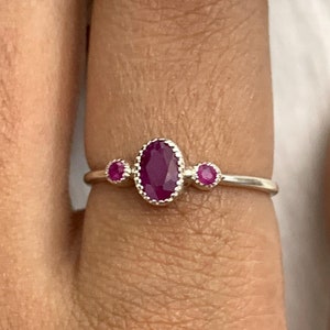 May include: A silver ring featuring an oval-shaped ruby gemstone in the center, flanked by two smaller round rubies. The ring's band is thin and delicate, showcasing the vibrant red gemstones. The ring is a piece of jewelry.