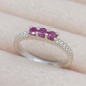 May include: A silver ring featuring three oval, deep red gemstones set in a row. The band has a textured, dotted design. The ring is displayed on a light pink surface, showcasing its elegant design.