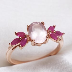 May include: Rose gold ring featuring an oval, light pink gemstone center, flanked by clusters of smaller, vibrant pink gemstones. The ring's band is a delicate rose gold, and the gemstones are set in a floral-inspired design.