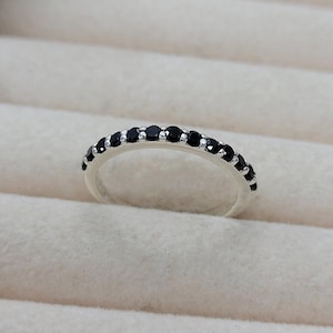 Può includere: Un anello d'argento con una fila di piccole pietre nere rotonde. L'anello è posto su uno sfondo bianco sporco, evidenziando il contrasto tra il metallo lucido e le pietre scure. Un gioiello delicato.