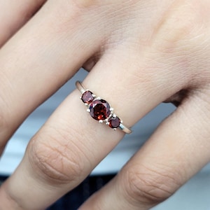 May include: A silver ring with three red gemstones. The ring is on a person's finger.