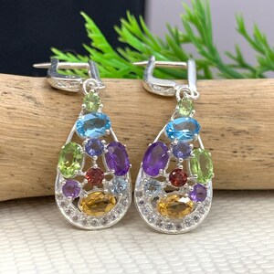 May include: A pair of silver dangle earrings in a teardrop shape. The earrings are decorated with a variety of colourful gemstones, including amethyst, peridot, topaz, and citrine.