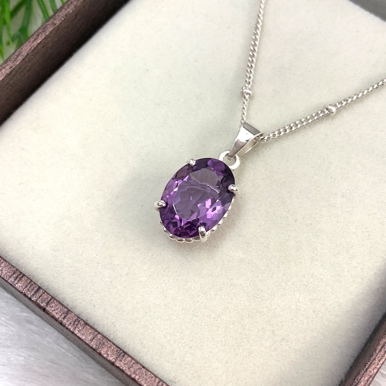Pode incluir: Um colar de corrente de prata com um pingente oval de pedra preciosa roxa. O pingente &eacute; engastado em uma moldura de prata com uma borda decorativa.